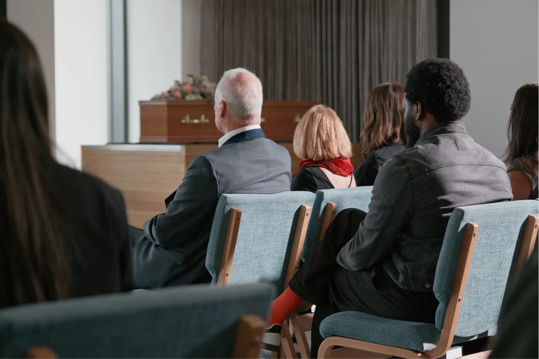 Mourners seated on chairs face a wooden coffin at the front of a light, modern chapel.