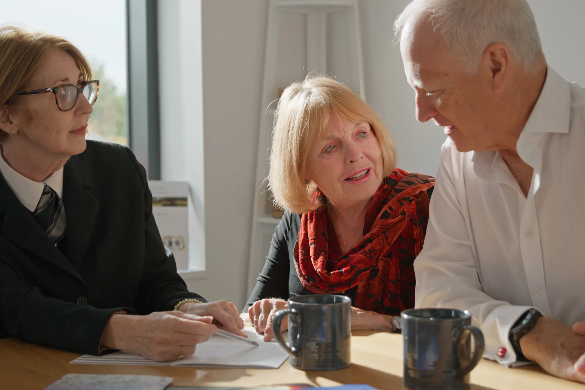 A funeral arranger in a dark suit takes notes with an older couple at a table.