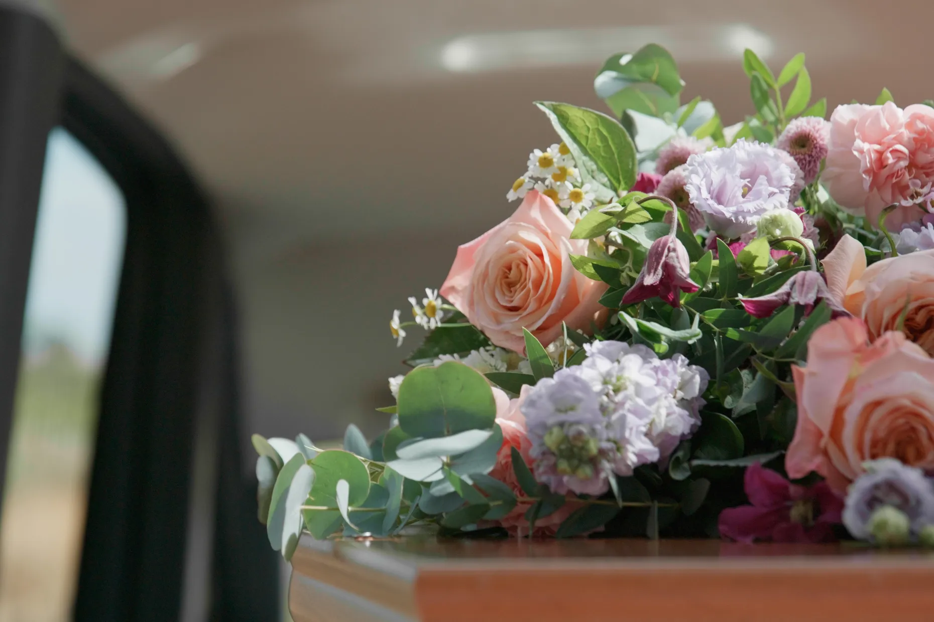 A wooden coffin inside a hearse, topped with a spray of peach roses and purple flowers.