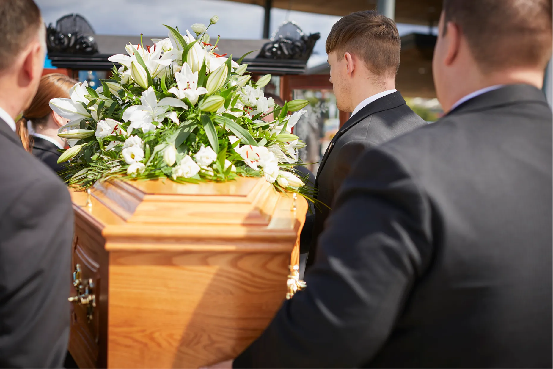 Bearers in dark suits carrying a light oak coffin topped with white lilies.