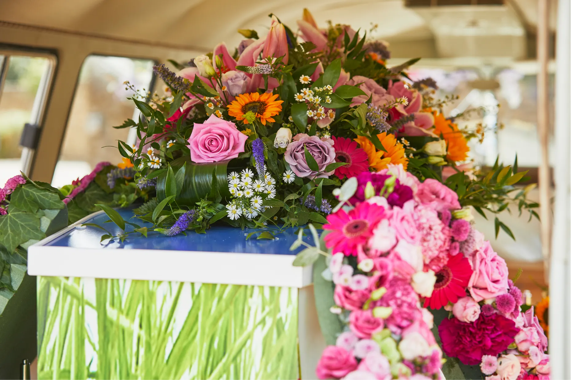 Bright roses, gerberas and sunflowers on a blue coffin, seen inside a vehicle.