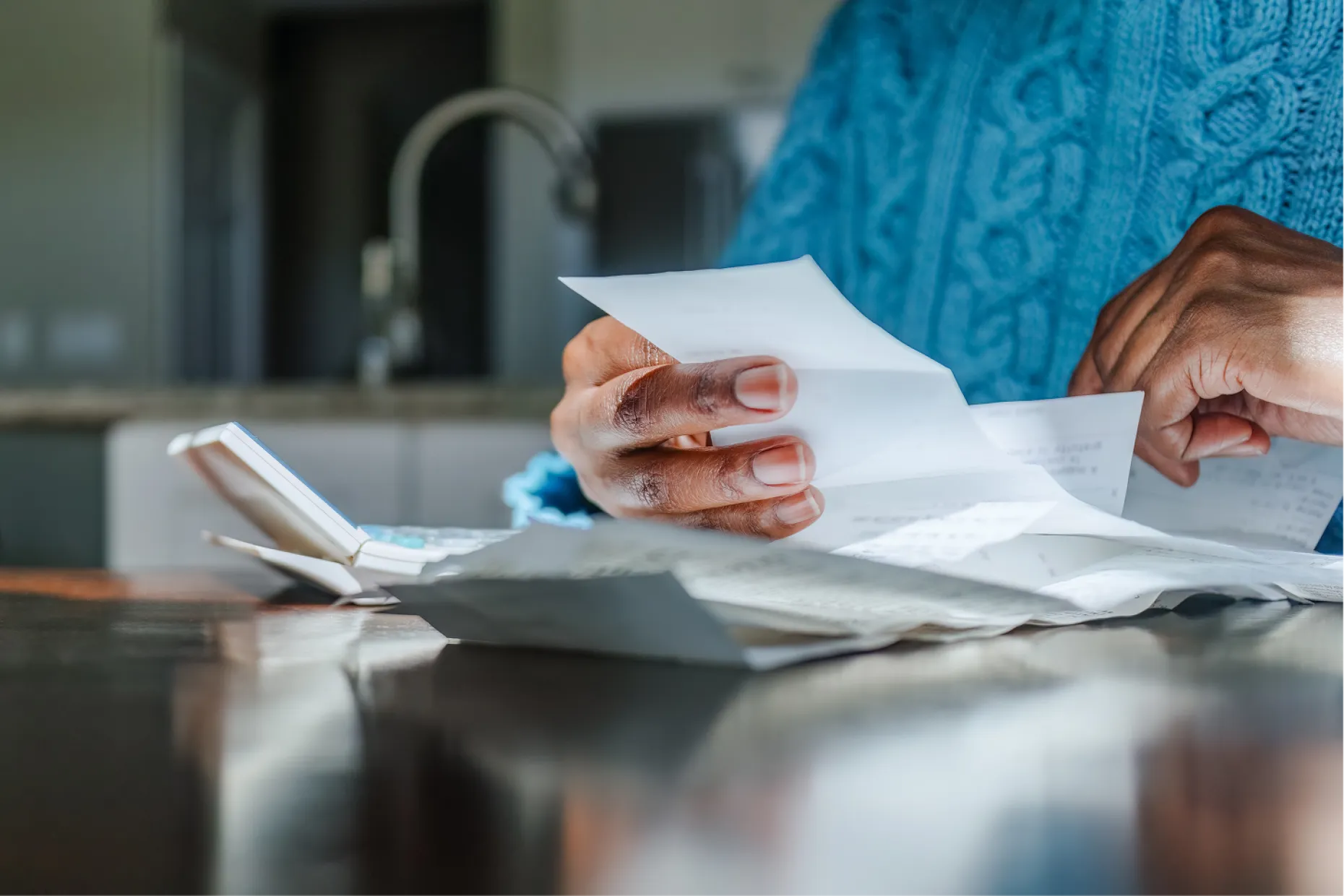 A person in a blue jumper reads a letter at a kitchen worktop, papers alongside.